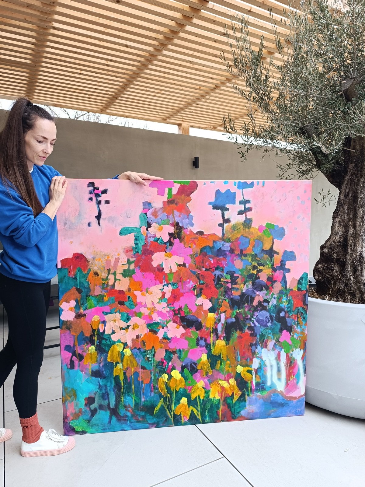 Autorka s čtvercovým abstraktním květinovým obrazem, růžové nebe a barevné květiny v živé kompozici